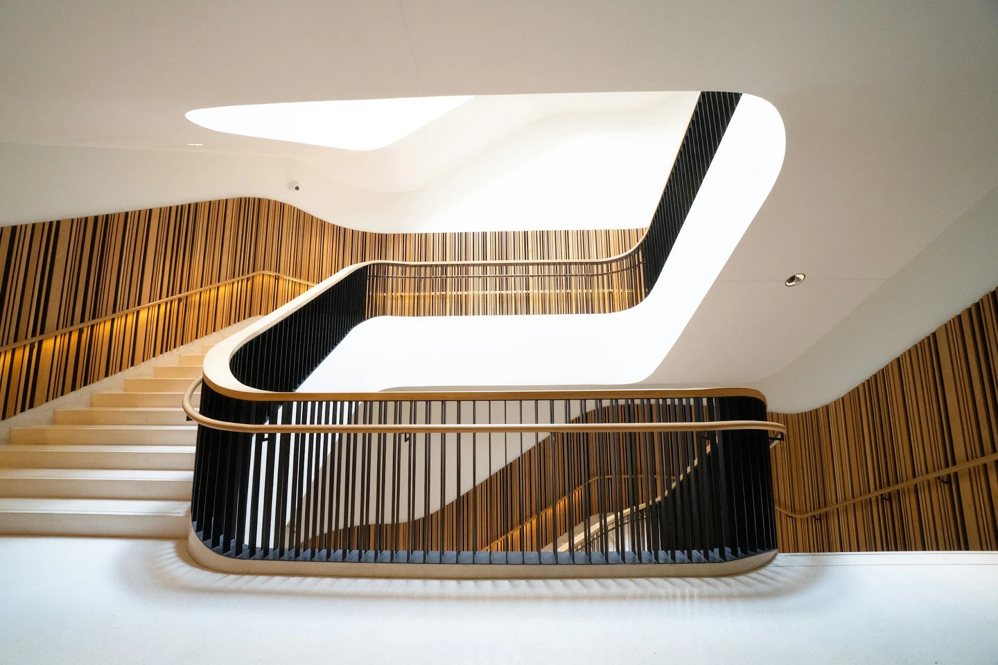 A winding staircase with black railings and warm wood-slat walls, lit from a skylight